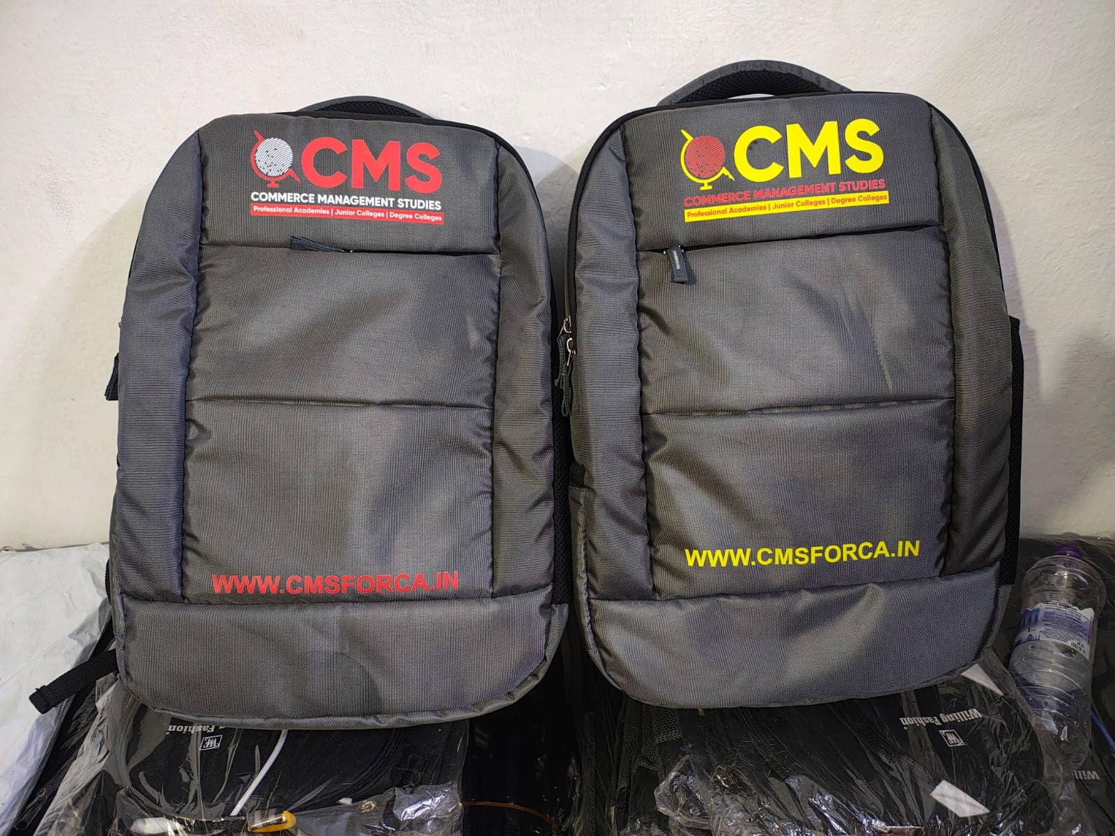 College backpacks printed with a customer's logo, standing on packed stock at the ABM Bags unit in Hyderabad